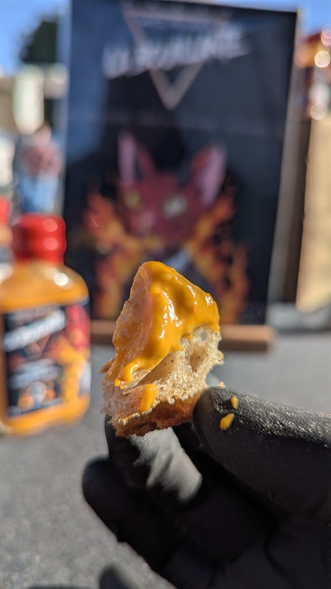 Une main gantée tient un morceau de pain couvert de sauce orange, devant un flacon flou et une illustration encadrée du chat.